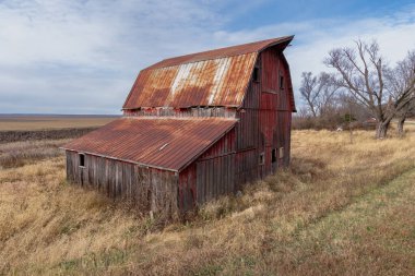Paslı metal çatılı, çürüyen, tarihi bir ahşap ahşap ahır, parçalı bulutlu bir gökyüzünün altında, uzun, kuru çayır otlarının arasında, kırsal terk edilmişliğin ve Orta Batı 'nın enginliğinin görüntüsünü yakalıyor..