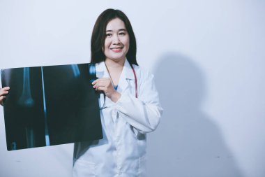 Tesettürlü Asyalı Müslüman doktor, boynuna kulaklık takıyor. Beyaz arka planda, elinde röntgen filmi tutarak güvenle ayakta duruyor..