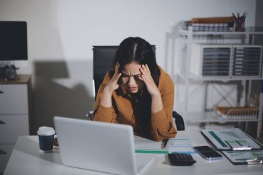 Bir kadın muhasebeci, ofis masasında dizüstü bilgisayarı üzerinde çalışırken bile başı ağrıyacak kadar belgelerle çalışmaktan stres altındaydı..