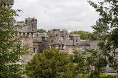 A grand, old stone structure stands majestically amid tall trees. This historic site features intricate details and round windows, creating an enchanting atmosphere on a cloudy day.