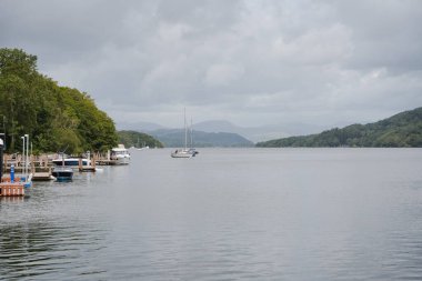Boats gently float on the lake as thick clouds hang overhead. Lush green trees line the shore, and distant mountains create a serene backdrop, showcasing a peaceful morning scene.