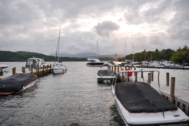 Boats are anchored at a calm marina, surrounded by lush green hills under a cloudy twilight sky. The water reflects the overcast ambiance as evening approaches.