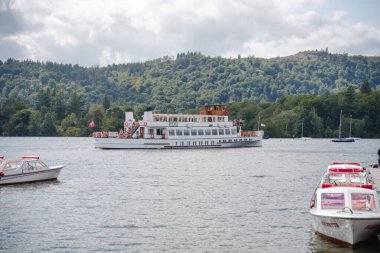 A large cruise boat glides smoothly across a serene lake. Smaller boats are anchored nearby as vibrant green hills rise in the background, creating a tranquil afternoon atmosphere.