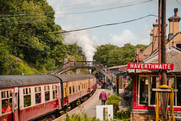 A vintage steam train pulls into Havertwaite station on a sunny day. Passengers eagerly await their journey as steam billows into the sky above the lush greenery.