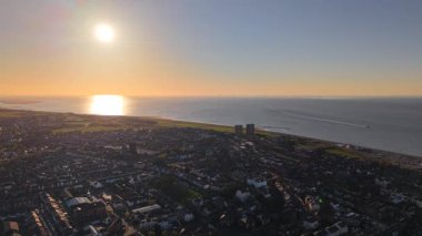 A breathtaking sunset casts vibrant colors over a coastal town. The sun reflects on the water, illuminating the beach and the distant wind turbines on the horizon.