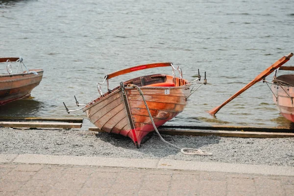 İki küçük renkli tekne çakıl taşlı bir sahil boyunca yanaşmış, bir tanesi hafifçe karaya çekilmiş. Temiz su güneşin altında parlıyor ve sakin bir atmosfer yaratıyor..