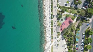 Bu çarpıcı hava görüntüsü turkuaz suların yanında bir sahil parkını yakalıyor. İnsanlar yol boyunca araçlar park halindeyken gezinti alanındaki ve yakınlardaki oyun alanındaki açık hava aktivitelerini seviyorlar..