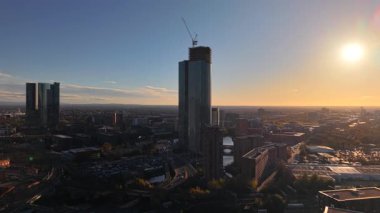 Manchester 'da inşaat halindeki yüksek bir bina belirgin bir şekilde duruyor. Güneş batıyor, şehrin her tarafına sıcak bir ışık saçıyor, modern ve geleneksel mimarinin bir karışımını ortaya çıkarıyor..