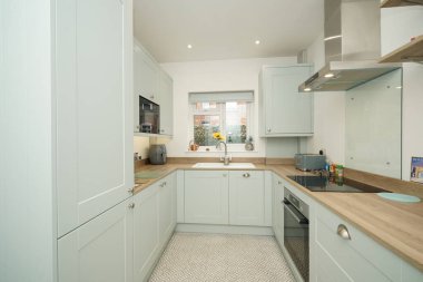 Hafif dolapları ve ahşap tezgahları olan temiz bir mutfak lavabo, soba ve pencereyi gösteriyor. Güneş ışığı odaya girer. Eşyalar düzgünce tezgahlara yerleştiriliyor..