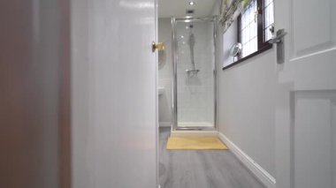 Temiz bir banyo, cam bir duş ve yerde bir paspas gösterir. Pencereden içeri ışık giriyor ve sabahları davet edici bir alan yaratıyor..