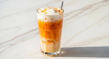 A tall glass of vibrant orange Thai iced tea, layered with creamy milk and ice, served on a marble surface with a spoon.