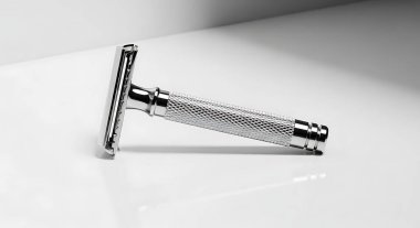 Beyaz bir yüzeyde duran parlak krom güvenlik jiletini yakından çek. Jilet keskin bir odak noktasında, karmaşık dizaynını ve metalik parıltısını vurguluyor. Arka plan temiz, minimalist beyaz, jiletleri vurguluyor..