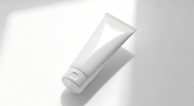 Cilt bakım ürünleri veya güzellik ürünleri için el değmemiş beyaz bir kozmetik tüpü temiz beyaz bir yüzeyde uzanır ve yumuşak, doğal gölgeler bırakır. Görüntü minimalist ve temiz bir estetiği vurgular, ürünlerin basitliğini ve zarafetini vurgular.