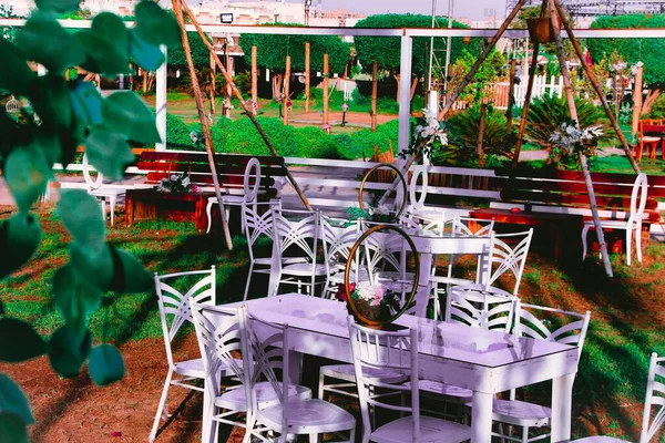 Beyaz mobilyaları ve çiçekli süslemeleri olan açık bir kafe. Masalar ve sandalyeler bahçe ortamında yemek için kurulur..