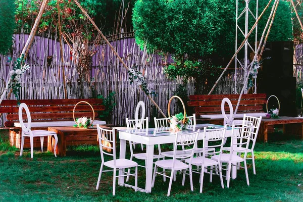 Yeşillik ve doğal ahşap yapılarla süslenmiş yeşil çimenlikte beyaz masalar ve sandalyelerle açık hava etkinliği..