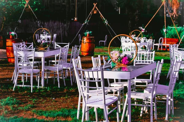 Açık hava etkinliği, beyaz masalar ve çimlerin üzerinde sandalyeler. Çiçek aranjmanları masaları süslüyor. Kırsal variller ve dekoratif yapılar arkada.