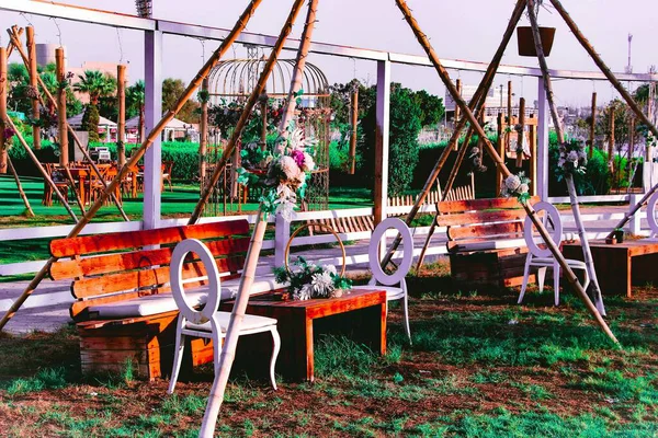 Çimenli bir alanda çadır benzeri bir yapının altında ahşap banklar, beyaz sandalyeler ve çiçek düzenlemeleri olan açık hava oturma alanı..