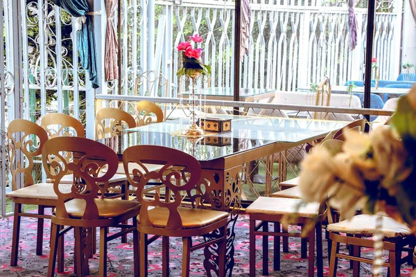 Süslü sandalyeleri ve cam kaplama masası olan, çiçeklerle ve kumaş perdelerle süslenmiş açık hava yemek salonu..