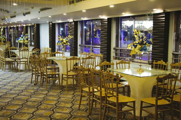 Masalar, sandalyeler, çiçekler ve desenli halılarla süslü bir etkinlik alanı. Geniş pencereler ışık girsin.