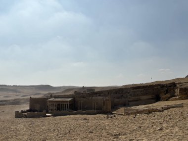 Parlak bir gökyüzünün altındaki çöl manzarasında antik kalıntılar. Kurak ortamda eski taş yapılar bulunur..