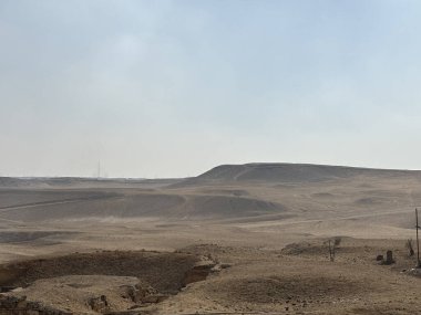 Puslu bir gökyüzünün altında yuvarlanan tepelerle çöl manzarası. Nadir bitki örtüsü. Ön planda bir hendek ya da hendek görünür..