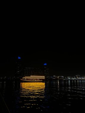 Bir tekne gece bir su kütlesinin üzerinde parladı. Binalar arka planda görülebilir.