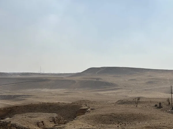 Puslu bir gökyüzünün altında yuvarlanan tepelerle çöl manzarası. Nadir bitki örtüsü. Ön planda bir hendek ya da hendek görünür..