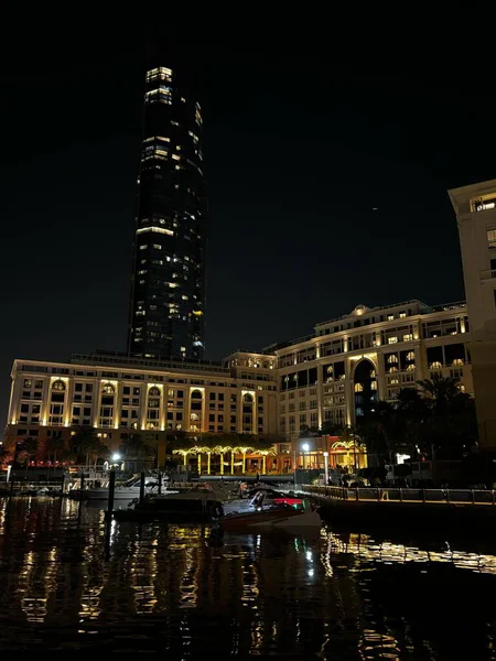 Uzun, aydınlık bir gökdelen ve bitişik süslemeli binanın gece görüşü suya yansıyor. Palmiye ağaçları ve tekneler görünür..