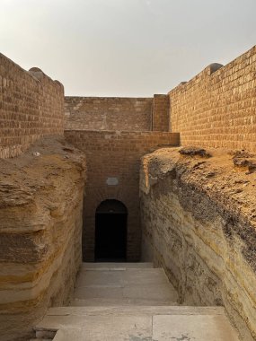 Bir yeraltı yapısına giriş, muhtemelen eski bir harabe ya da mezar. Taş duvarlar ve basamaklar karanlık bir geçide çıkar..