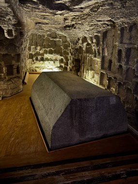 Yukarıdan aydınlatılmış, mezar benzeri bir ortamda karanlık taş lahit. Eserin etrafını oyulmuş taş duvarlar sarmış..