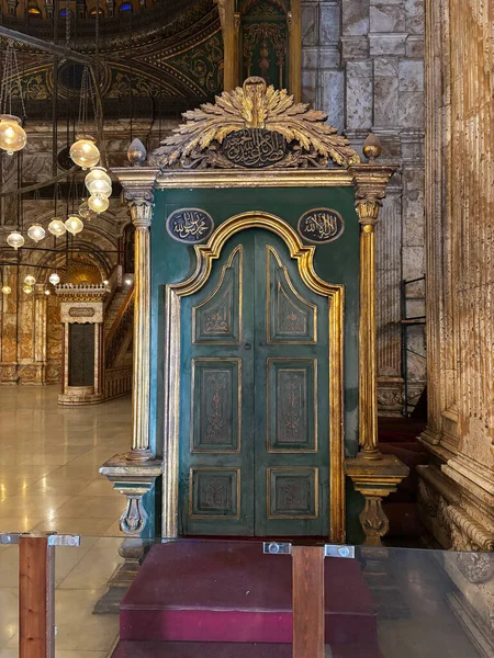 Bir binanın içindeki Arapça yazıyla süslenmiş yeşil & altın kapı. Arka planda asılı lambalar da görülüyor.