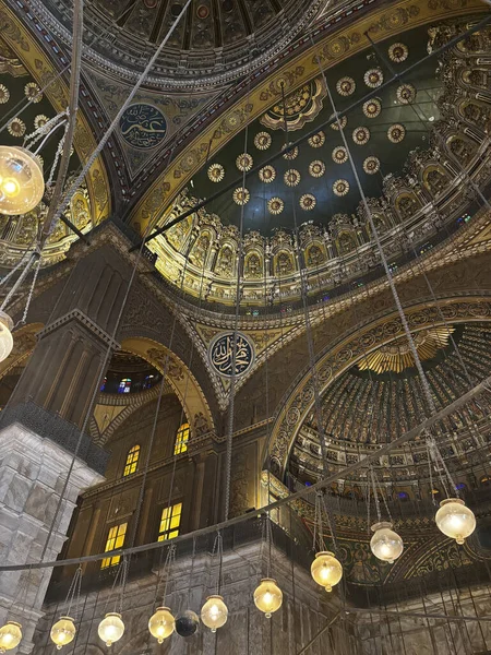 Bir caminin iç mekanının karmaşık mimari ayrıntıları, dekoratif tavanlar, kemerler ve asılı lambaların yer aldığı. Altın ve koyu yeşil renk şeması.