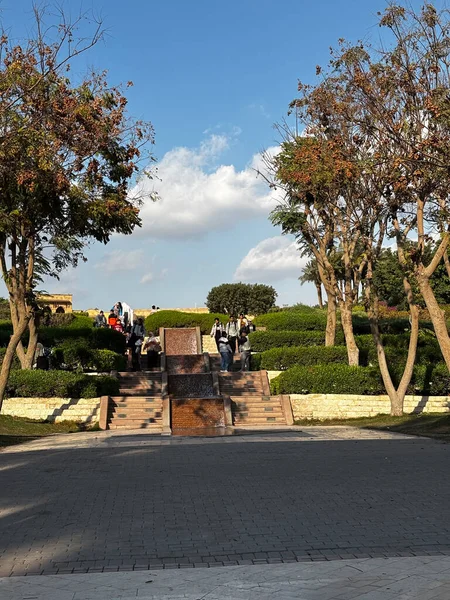 Katmanlı fıskiyeli, ağaçlı ve insanlı park sahnesi. Bulutlu mavi gökyüzü.