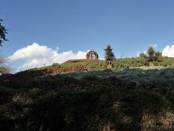 Mavi gökyüzünün altındaki çimenli bir tepenin tepesinde, dağınık bulutlu bir yapı. Yamaçta yeşillik ve bitki örtüsü var..