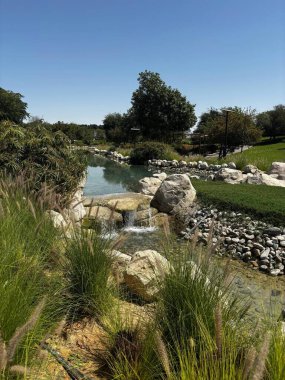 Açık mavi gökyüzünün altında küçük şelaleler, kayalar, çimenler ve yeşillikler olan manzaralı bir su yolu manzarası..