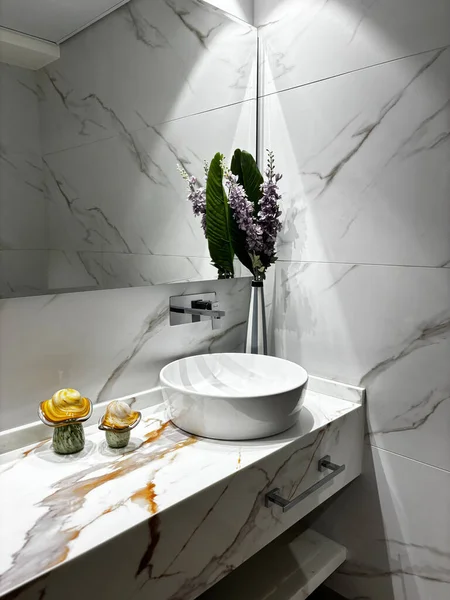 Mermer tezgahları ve duvarları olan modern bir banyo. Yuvarlak bir lavabo, dekoratif eşyalar ve bir vazo çiçekle tezgahın üzerinde duruyor..