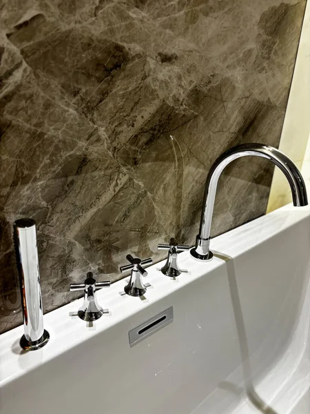 Pürüzsüz, modern bir banyo musluğunun mermer desenli bir duvara yakın çekimi. Cilalanmış krom eşyalar ve minimalist bir tasarım..