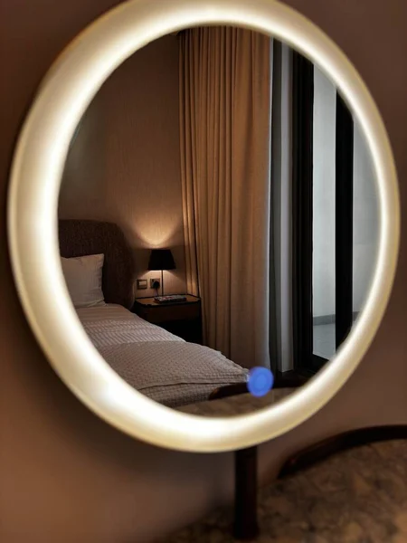 Yuvarlak, aydınlık bir ayna yatak odası sahnesini yansıtıyor yatak odası, komodin ve lambayla. Aynada sıcak bir parıltı var..