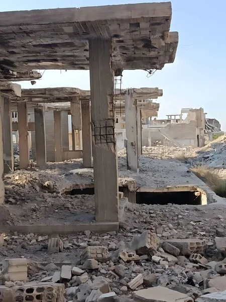 Yıkılmış bina. Beton sütunlar ve molozlar sahneyi dolduruyor. Bina harabe halinde, arka planda mavi bir gökyüzü görünüyor..