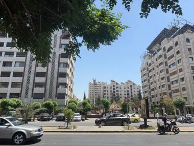 Şehrin tepetaklak manzarası yüksek binalar, arabalar ve ağaçlarla aydınlık bir gökyüzünün altında.