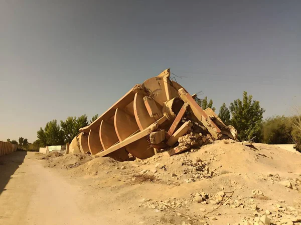 Büyük, harap bir yapı enkaz ve kir içinde ıssız ve çorak bir arazide yatıyor. Ağaçlar ufuk çizgisinde berrak bir gökyüzünün altında.