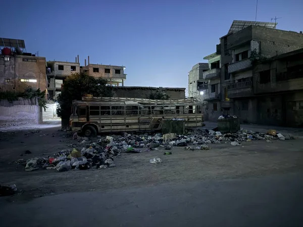 Şehir ortamında çöplerin arasında, mavi bir alacakaranlık gökyüzünün altındaki binaların arasında eski bir otobüsün ters dönmüş görüntüsü..