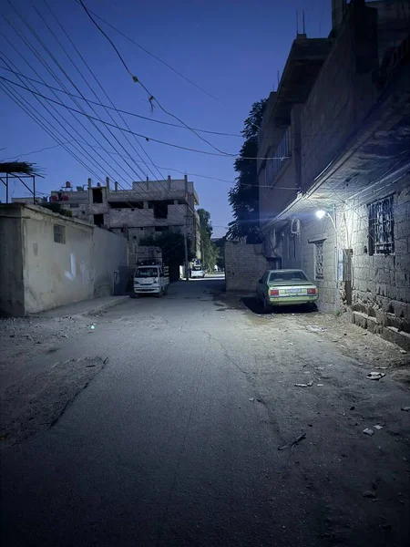 Alacakaranlıkta dar bir sokak. Binalar cadde boyunca park halindeki arabalarla ve kablolar görünür durumda..