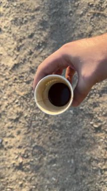İçinde az miktarda koyu sıvı bulunan bir kağıt bardağı tutan bir elin yakın çekimi, muhtemelen kahve, kum veya toprak zeminine karşı..