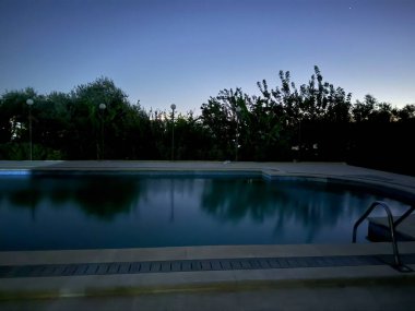 Alacakaranlıkta ya da şafakta yüzme havuzu, gökyüzünü ve çevresindeki yeşilliği yansıtıyor. Sakin ve sakin bir atmosfer.