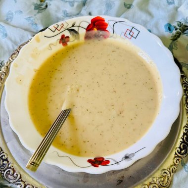 Süslü beyaz bir kâsede, gümüş tepside sunulan kremalı çorba ve çiçek desenli masa örtüsü..