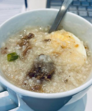 İçinde yumurta ve et olan bir kupa pirinç lapası, içinde kaşık olan bir tabak üzerinde oturuyor, ve arka planda kısmen görünen bir dizüstü bilgisayar klavyesi var..