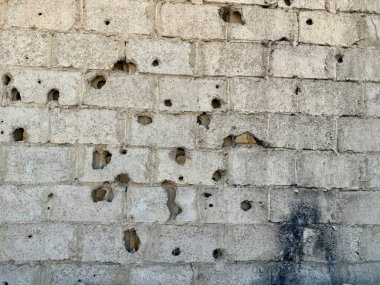 Beton blok duvarda çok sayıda delik ve hasar var. Gri bloklar eskimiş ve yıpranmış. Doku pürüzlü ve darbe belirtileri gösteriyor..
