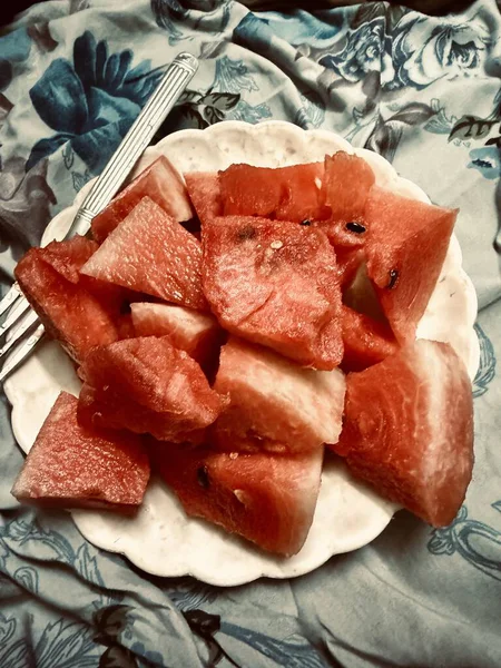 Bir çatalla bir tabakta doğranmış karpuz, mavi çiçekli bir yüzey üzerinde duruyor. Parlak ve yaz.