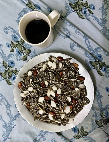 Bir kupa kahvenin ve çiçek desenli bir yüzeye bir tabak karışık ayçekirdeği ve fındık..